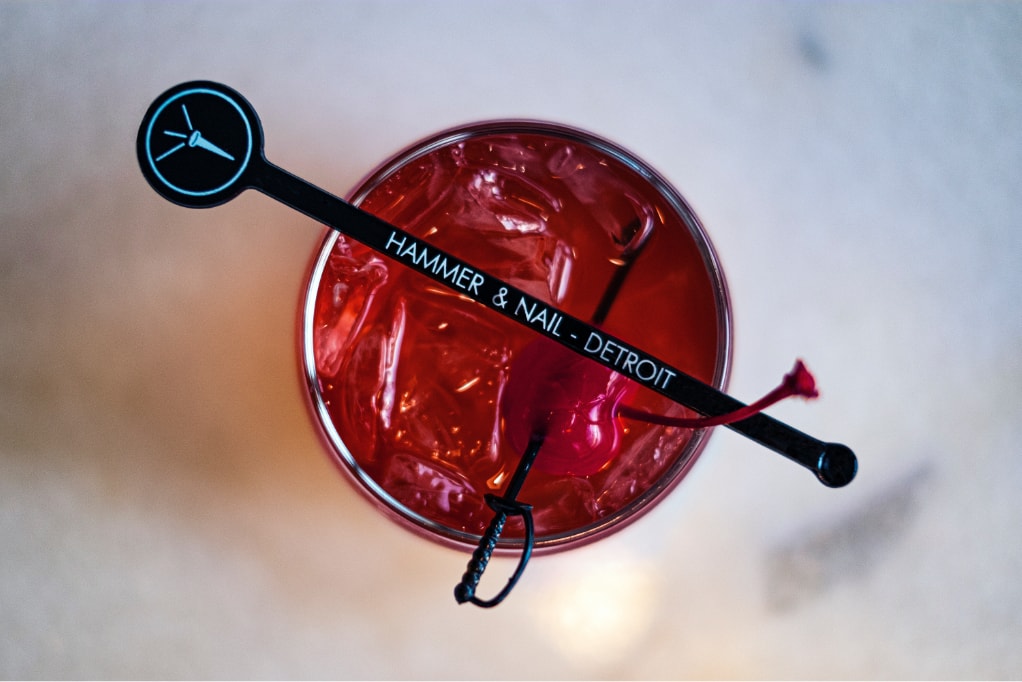 A deep red Old Fashioned cocktail, shot from above, with a branded Hammer and Nail cocktail stirrer laying over the glass