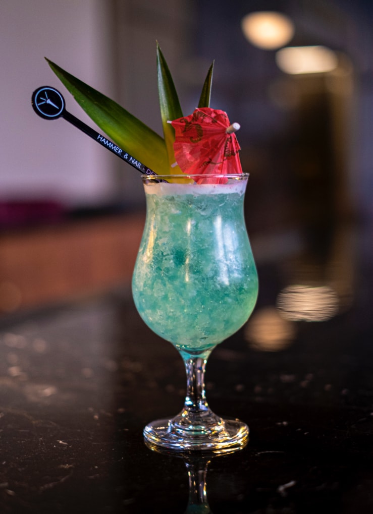 The bright Blue Hawaii cocktail sits on a black marble bar top, dressed up in a hurricane glass with a cocktail umbrella and pineapple leaves