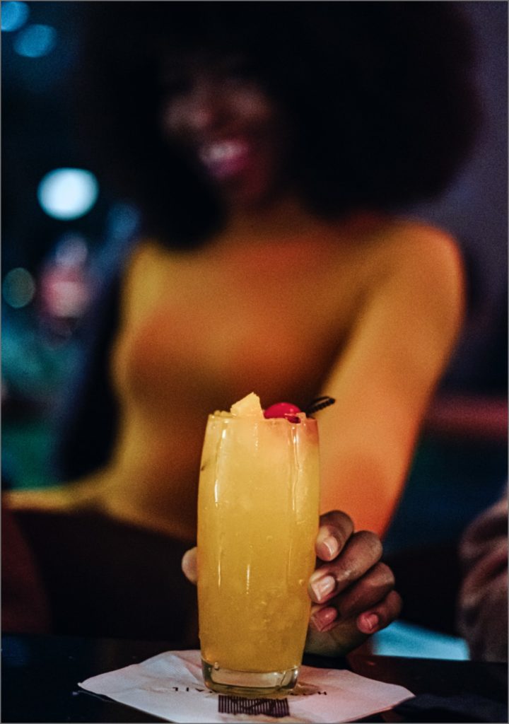 A woman in a yellow shirt holds an orange-yellow Wallbanger cockatail