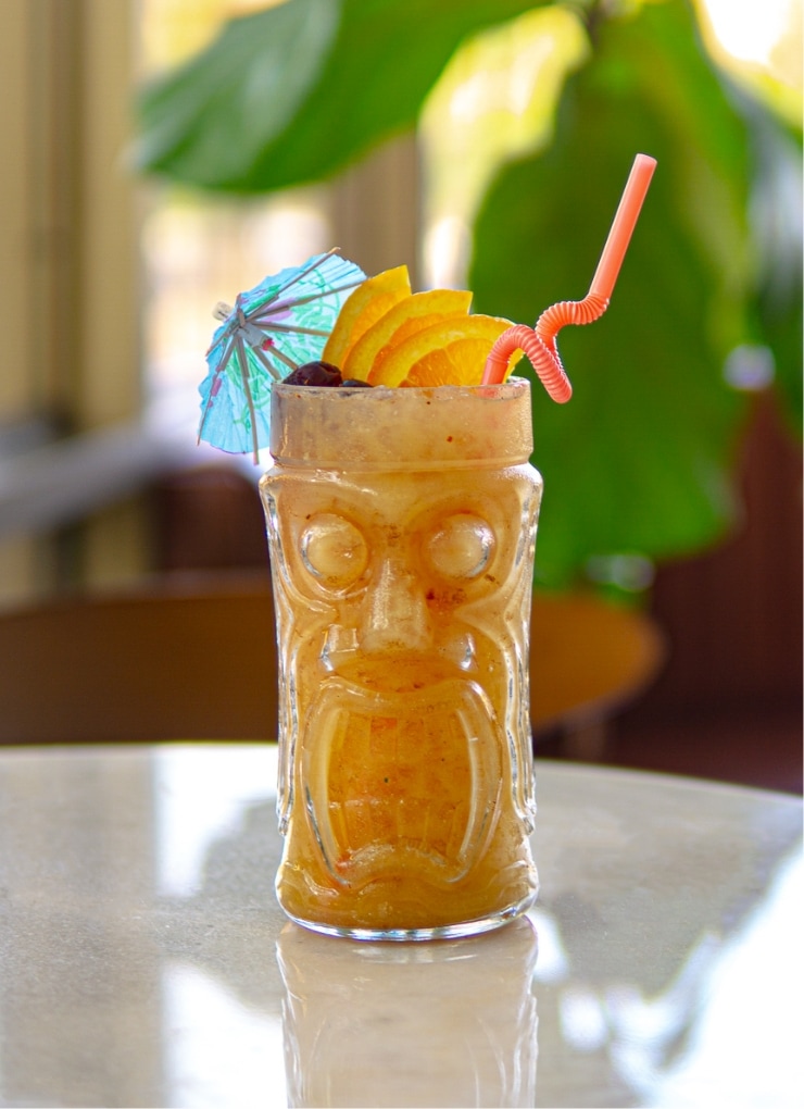 A tiki cocktail sits on a white marble table, dressed up in a tiki mug with a cocktail umbrella, cherries, and orange slices