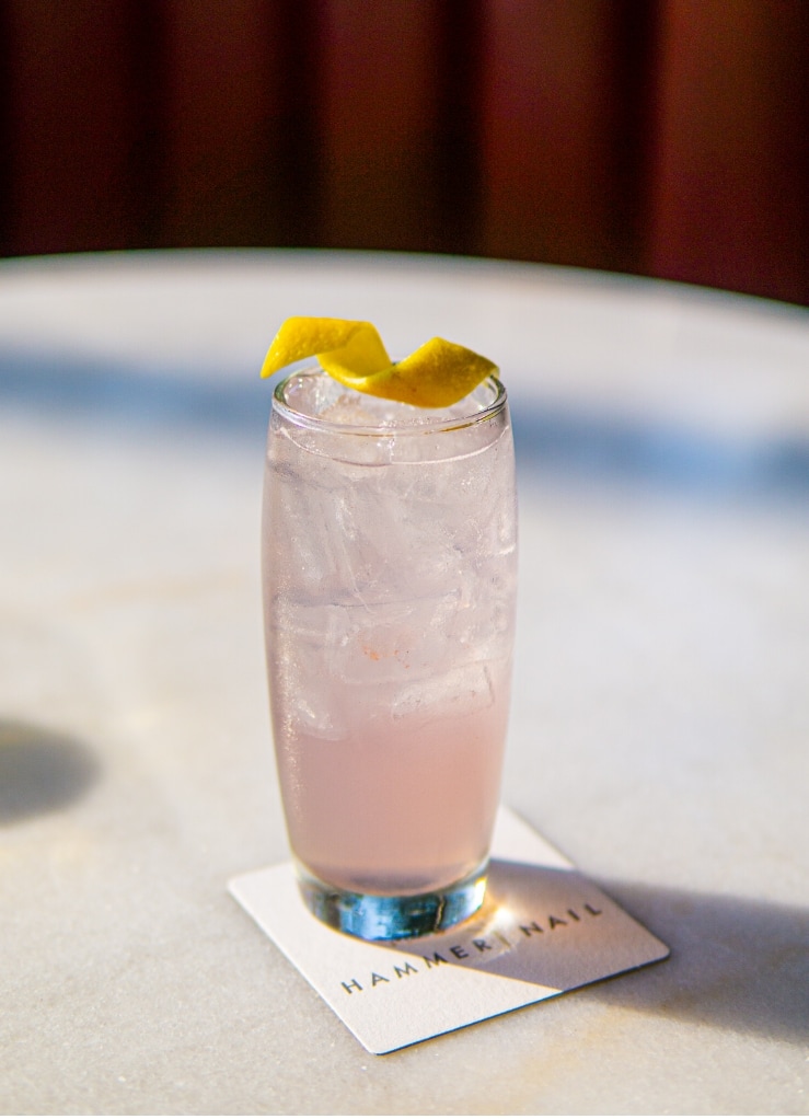 A pink cocktail sits on a Hammer and Nail coaster, topped with a lemon twist